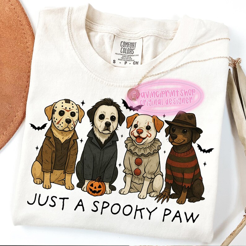 May include: Cream-colored t-shirt featuring four cartoon dogs dressed as horror movie characters. The phrase "JUST A SPOOKY PAW" is printed below the dogs. A gold necklace and a pink tag are also visible.