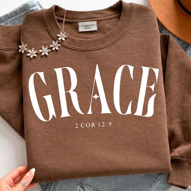 K&ouml;nnte beinhalten: Ein braunes Sweatshirt mit dem Wort "GRACE" in gro&szlig;en wei&szlig;en Buchstaben. Unter dem Wort steht der Text "2 COR 12:9". Eine Blumenkette ist &uuml;ber dem Sweatshirt drapiert.