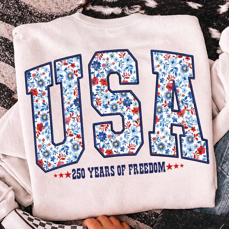 May include: Cream-colored sweatshirt with the word "USA" in large, block letters. The letters are filled with a floral pattern of blue and red flowers. Below the letters is the text "250 YEARS OF FREEDOM" with stars.