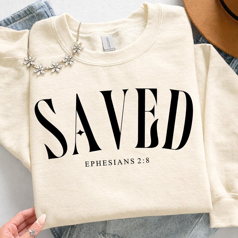 K&ouml;nnte beinhalten: Cremefarbenes Sweatshirt mit dem Wort "SAVED" in gro&szlig;en, fetten, schwarzen Buchstaben. Darunter steht der Text "EPHESIANS 2:8". Eine Blumenkette und ein brauner Hut sind ebenfalls zu sehen.