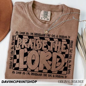 May include: A brown t-shirt with a black and white checkered design and the text "Praise The Lord" in black lettering. The text "As Long As I'm Breathing I've Got a Reason To" is repeated at the top and bottom of the design.