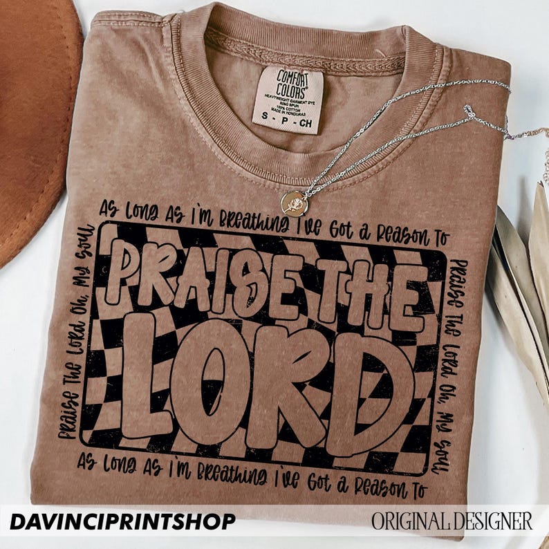 May include: A brown t-shirt with a black and white checkered design and the text "Praise The Lord" in black lettering. The text "As Long As I'm Breathing I've Got a Reason To" is repeated at the top and bottom of the design.