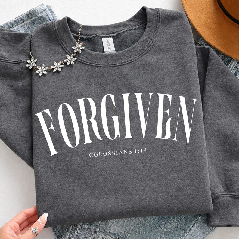 K&ouml;nnte beinhalten: Dunkelgraues Sweatshirt mit dem Wort "FORGIVEN" in gro&szlig;en wei&szlig;en Buchstaben. Darunter steht der Text "COLOSSER 1:14". Eine silberne Blumenkette liegt auf dem Sweatshirt.