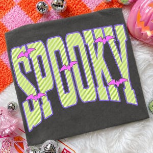 May include: A dark gray t-shirt featuring the word SPOOKY in large, lime green and white striped letters with a purple outline. The design includes small, bright pink bat silhouettes positioned between and around the letters of the text.