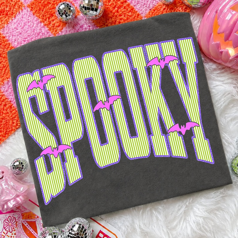 May include: A dark gray t-shirt featuring the word SPOOKY in large, lime green and white striped letters with a purple outline. The design includes small, bright pink bat silhouettes positioned between and around the letters of the text.