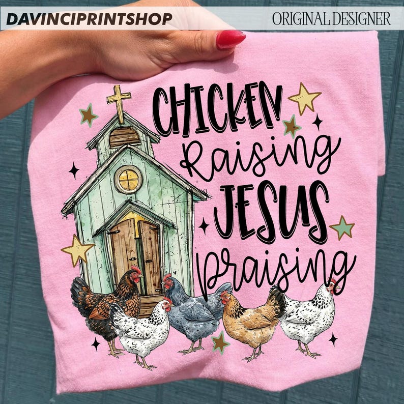 Peut inclure: T-shirt rose avec un motif d'&eacute;glise, de poulets et les mots "Chicken Raising Jesus Praising". Le design comprend des &eacute;toiles et une croix. Le t-shirt est tenu par une personne.