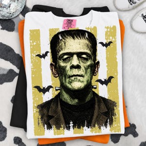 May include: A white t-shirt featuring a graphic of Frankenstein's monster in shades of green and black. The design includes vertical yellow distressed stripes and black bat silhouettes. A pink Comfort Colors brand tag is visible at the neckline.