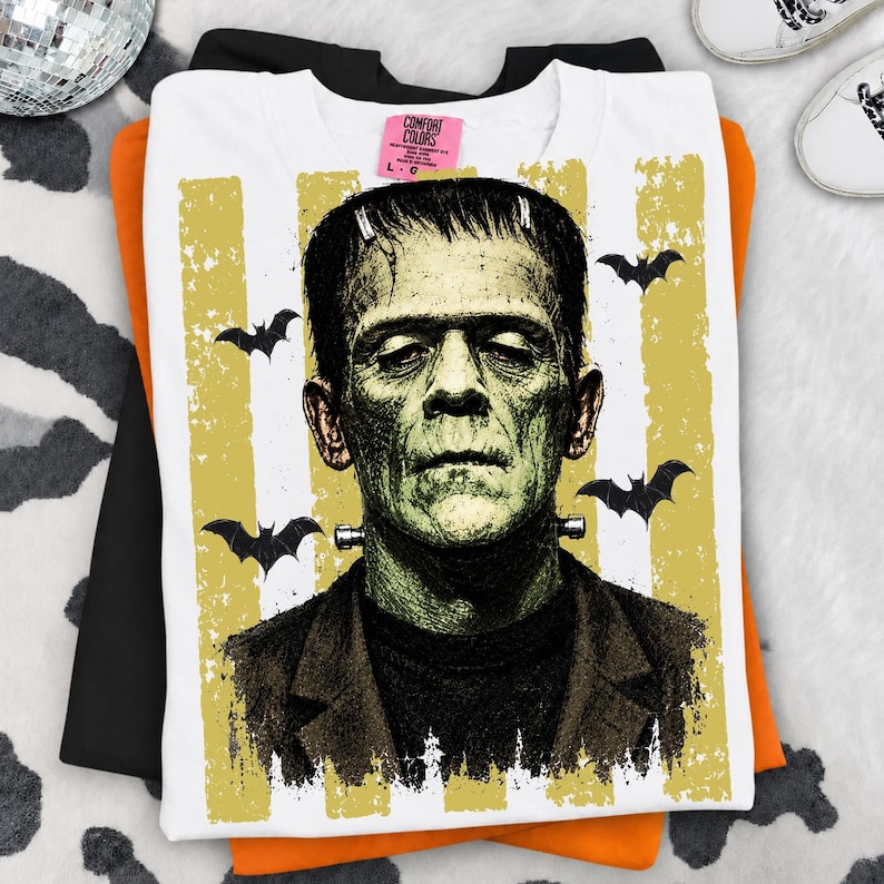 May include: A white t-shirt featuring a graphic of Frankenstein's monster in shades of green and black. The design includes vertical yellow distressed stripes and black bat silhouettes. A pink Comfort Colors brand tag is visible at the neckline.