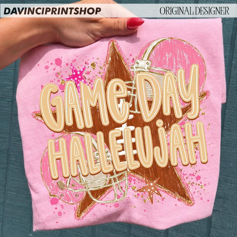 May include: A light pink t-shirt featuring a graphic print of a brown star, two pink football helmets, and pink star accents. The text GAME DAY HALLELUJAH is printed in large, light tan letters with a white outline across the center of the design.