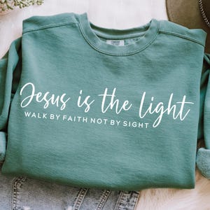 May include: Teal blue sweatshirt with the text "Jesus is the light" and the phrase "Walk by faith not by sight" in white lettering.