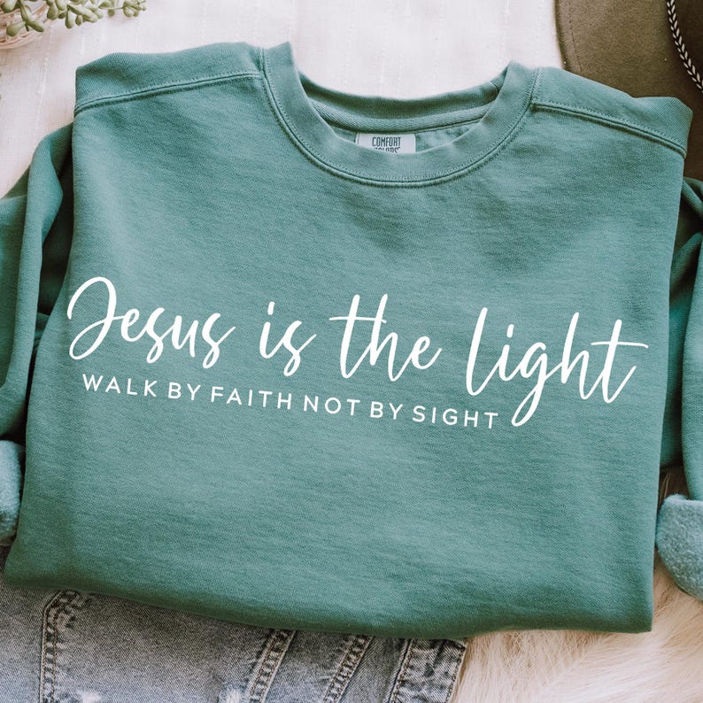 May include: Teal blue sweatshirt with the text "Jesus is the light" and the phrase "Walk by faith not by sight" in white lettering.
