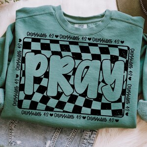 Op de afbeelding: Een turquoise sweatshirt met een zwart-wit geruit patroon en het woord "PRAY" in grote witte letters. Het patroon is een vervormd ruitpatroon met een vintage look. De tekst "Colossians 4:2" wordt herhaald rond de rand van het ruitpatroon.