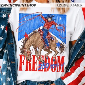 May include: White t-shirt featuring a cowboy riding a horse with a lasso, set against a blue and red backdrop with stars. The word "FREEDOM" is in red, with "250 years of" in blue below. The shirt has the text "DAVINCIPRINTSHOP" and "ORIGINAL DESIGNER".