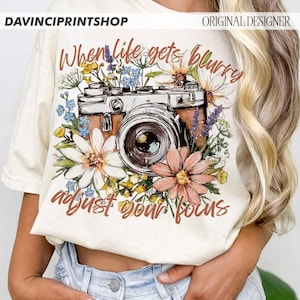May include: Cream-colored t-shirt featuring a vintage camera design surrounded by colorful flowers. The text "When life gets blurry adjust your focus" is written in brown script. The shirt has a relaxed fit.