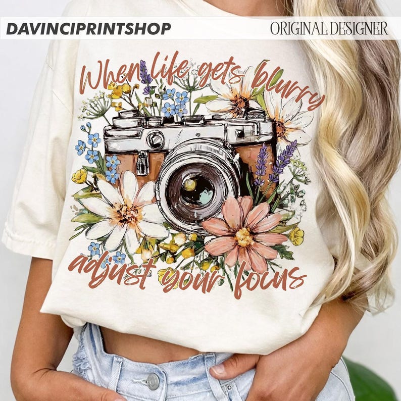 May include: Cream-colored t-shirt featuring a vintage camera design surrounded by colorful flowers. The text "When life gets blurry adjust your focus" is written in brown script. The shirt has a relaxed fit.