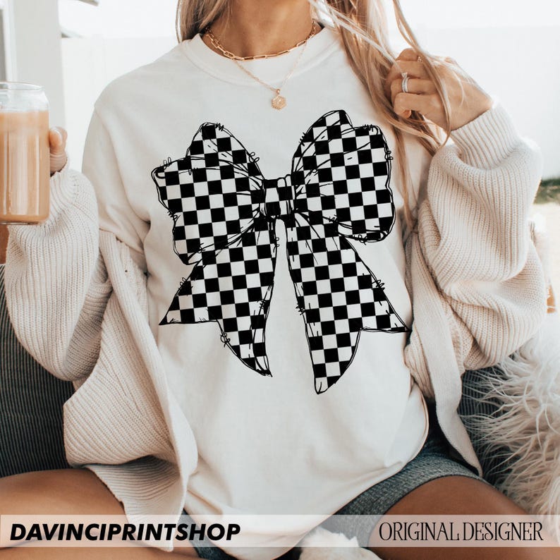 May include: Cream-colored sweatshirt featuring a large black and white checkered bow graphic. The text "DAVINCIPRINTSHOP" is at the bottom left, and "ORIGINAL DESIGNER" is at the bottom right. The sweatshirt has long sleeves.
