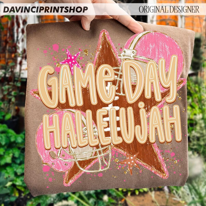 May include: A brown t-shirt featuring a graphic print of a large brown star, two pink football helmets, and pink star accents. The text "GAME DAY HALLELUJAH" is printed in large, light tan letters across the center of the graphic design.