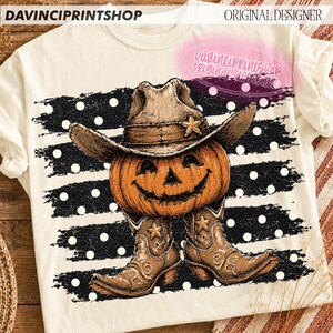K&ouml;nnte beinhalten: Ein cremefarbenes T-Shirt mit einer Grafik eines K&uuml;rbisses, der einen braunen Cowboyhut und braune Cowboystiefel tr&auml;gt. Das Motiv ist vor einem Hintergrund aus schwarzen horizontalen Pinselstrichen mit wei&szlig;en Punkten und dem Text DAVINCIPRINTSHOP.
