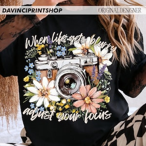 May include: Black t-shirt featuring a vintage camera design surrounded by colorful flowers. The text "When life gets blurry, adjust your focus" is written in white script. The shirt has the text "DAVINCIPRINTSHOP" and "ORIGINAL DESIGNER" at the top.