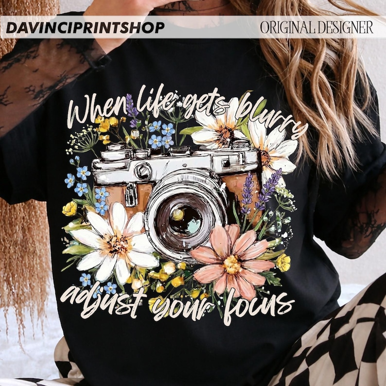 May include: Black t-shirt featuring a vintage camera design surrounded by colorful flowers. The text "When life gets blurry, adjust your focus" is written in white script. The shirt has the text "DAVINCIPRINTSHOP" and "ORIGINAL DESIGNER" at the top.