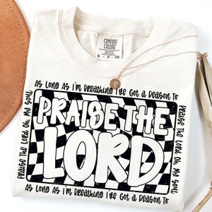 May include: A beige t-shirt with a black and white checkered design and the text "Praise The Lord" in a bold, retro font. The text "As Long As I'm Breathing I've Got a Reason To" is repeated at the top and bottom of the design.