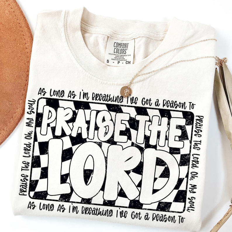 May include: A beige t-shirt with a black and white checkered design and the text "Praise The Lord" in a bold, retro font. The text "As Long As I'm Breathing I've Got a Reason To" is repeated at the top and bottom of the design.