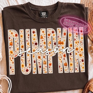 May include: A dark brown t-shirt featuring the word PUMPKIN in large block letters filled with a floral and pumpkin pattern, outlined in white faux pearls. The word season is written in white cursive script across the center of the letters.