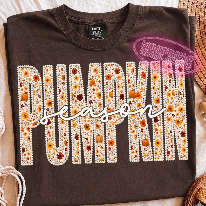 May include: A dark brown t-shirt featuring the word PUMPKIN in large block letters filled with a floral and pumpkin pattern, outlined in white faux pearls. The word season is written in white cursive script across the center of the letters.