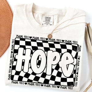 Op de afbeelding: Een cr&egrave;mekleurig T-shirt met een zwart-wit geruit patroon en het woord "HOPE" in een retro-lettertype. De tekst "Psalm 130:5" wordt herhaald rond het geruite patroon.