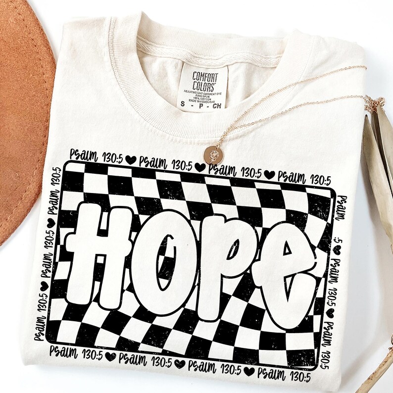 Op de afbeelding: Een cr&egrave;mekleurig T-shirt met een zwart-wit geruit patroon en het woord "HOPE" in een retro-lettertype. De tekst "Psalm 130:5" wordt herhaald rond het geruite patroon.