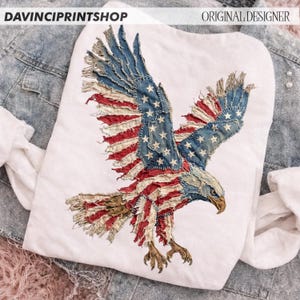 Pu&ograve; includere: T-shirt bianca con un design di aquila della bandiera americana invecchiata. Le ali dell'aquila sono spiegate, mostrando le strisce rosse e bianche e le stelle blu. I testi "DAVINCIPRINTSHOP" e "ORIGINAL DESIGNER" sono visibili.