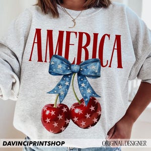 May include: A grey sweatshirt with the word "AMERICA" printed in red letters. Below the word is a blue bow with white stars, and two red cherries with white stars.