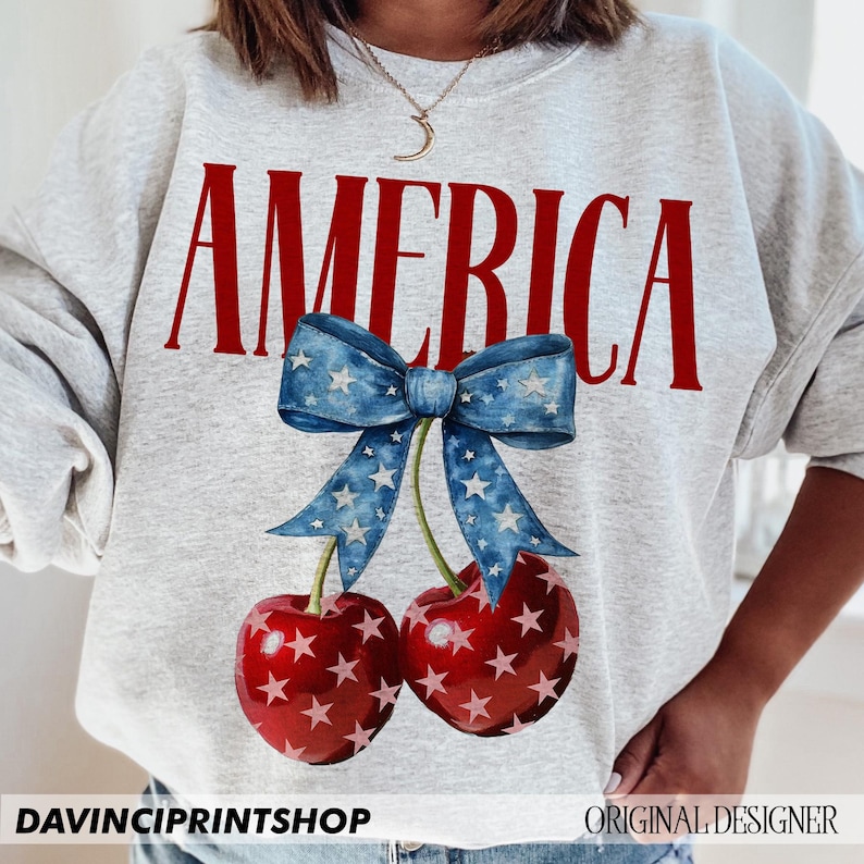 May include: A grey sweatshirt with the word "AMERICA" printed in red letters. Below the word is a blue bow with white stars, and two red cherries with white stars.