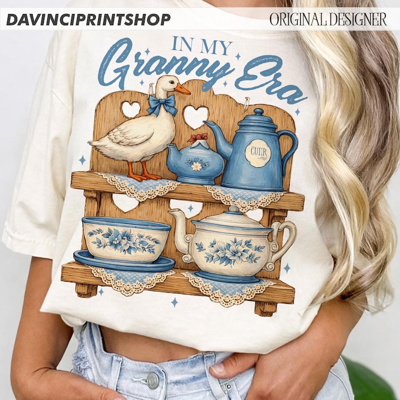 Puede incluir: Camiseta de color crema con una ilustraci&oacute;n caprichosa de un pato, teteras y tazas en una estanter&iacute;a de madera. La frase "IN MY Granny Era" est&aacute; impresa en azul sobre el dise&ntilde;o. El dise&ntilde;o incluye patrones florales azules y blancos.