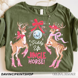 May include: An olive green t-shirt with a Christmas-themed design. It features two deer in pink cowboy boots and scarves, with the text "Giddy Up Jingle Horse!". A disco ball and bow are also included. The shirt has the text "DAVINCIPRINTSHOP".