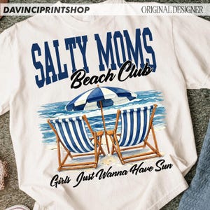 Op de afbeelding: Wit T-shirt met de tekst "Salty Moms Beach Club" in blauw en zwart. Het shirt heeft een strandsc&egrave;ne met twee blauw-wit gestreepte strandstoelen, een parasol en de woorden "Girls Just Wanna Have Sun".
