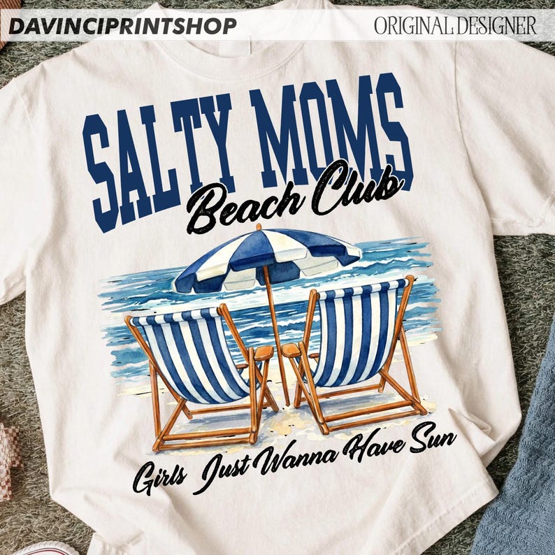 Op de afbeelding: Wit T-shirt met de tekst "Salty Moms Beach Club" in blauw en zwart. Het shirt heeft een strandsc&egrave;ne met twee blauw-wit gestreepte strandstoelen, een parasol en de woorden "Girls Just Wanna Have Sun".