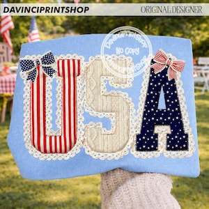 May include: A light blue decorative item with the letters "USA" in red, white, and blue. The "U" has red and white stripes, the "S" is a cable knit design, and the "A" is navy blue with white stars. Each letter is outlined with white lace and topped with a bow.