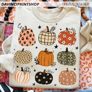May include: A beige crewneck sweatshirt featuring a graphic print of nine pumpkins with various patterns including leopard print, plaid, polka dots, floral, and heart motifs, interspersed with small black stars and vine illustrations.
