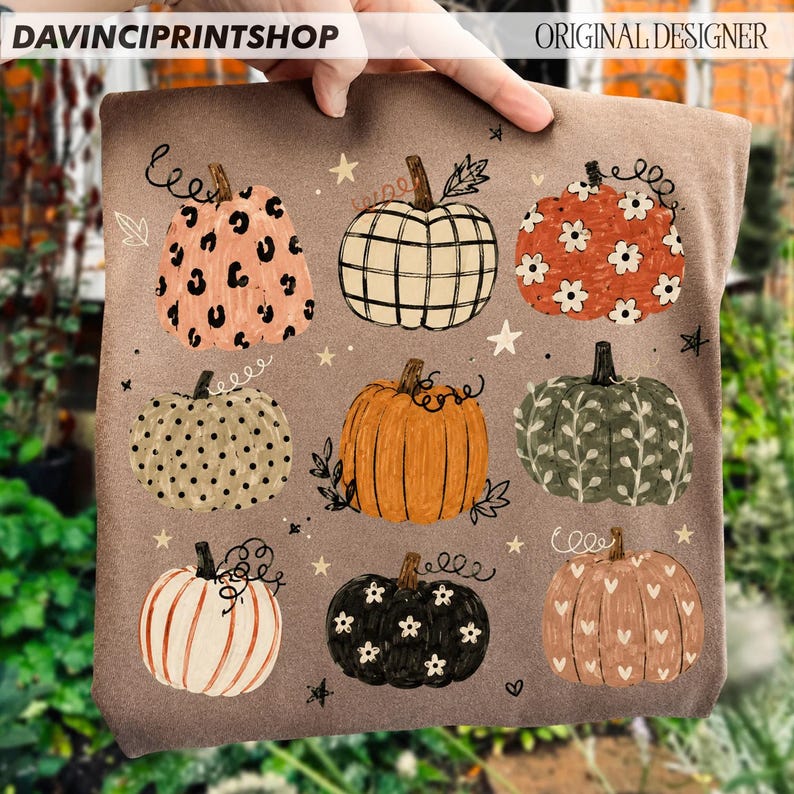 May include: A brown t-shirt featuring a graphic print of nine pumpkins in various patterns including leopard print, plaid, floral, polka dot, and heart designs, accented with small stars and swirling vine illustrations across the fabric.