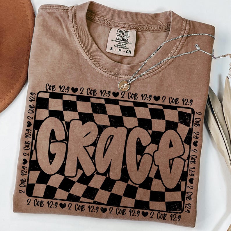 Op de afbeelding: Een bruin T-shirt met een zwart-wit geruit patroon en het woord "Grace" in een groot, gestileerd lettertype. De tekst "2 Cor 12:9" wordt herhaald rond het geruite patroon.