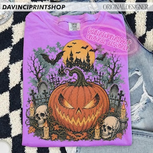 May include: A light purple t-shirt featuring a graphic print of a large orange jack-o'-lantern, two human skulls, and a dark silhouette of a haunted castle under a full moon. The design includes bare trees, tombstones, and bats in a spooky scene.