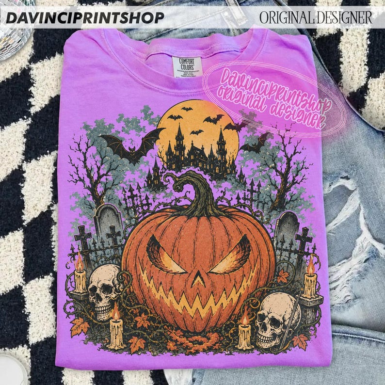 May include: A light purple t-shirt featuring a graphic print of a large orange jack-o'-lantern, two human skulls, and a dark silhouette of a haunted castle under a full moon. The design includes bare trees, tombstones, and bats in a spooky scene.