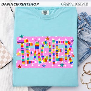 May include: A light blue short-sleeve t-shirt featuring a pink rectangular graphic with white polka dots. The word TEACHER is printed in large, multi-colored striped letters across the center, surrounded by small, colorful star icons.