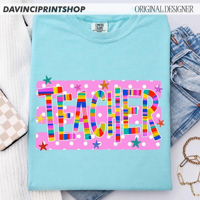 May include: A light blue short-sleeve t-shirt featuring a pink rectangular graphic with white polka dots. The word TEACHER is printed in large, multi-colored striped letters across the center, surrounded by small, colorful star icons.