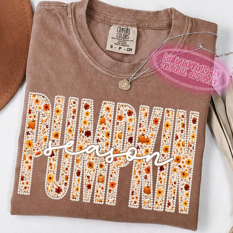 May include: A brown short-sleeved t-shirt featuring the word PUMPKIN in large block letters filled with a floral pattern of orange, yellow, and brown flowers and pumpkins, with the word season written in white cursive script across the center.