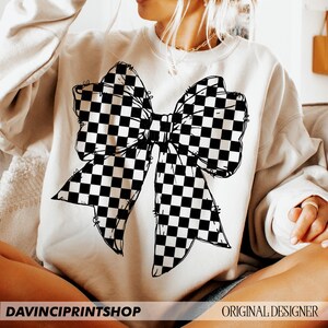May include: Cream-colored sweatshirt featuring a large black and white checkered bow graphic. The bow design is centered on the front of the shirt. The text "DAVINCIPRINTSHOP" is at the bottom left, and "ORIGINAL DESIGNER" is at the bottom right.