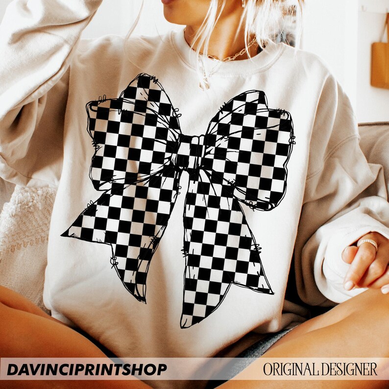 May include: Cream-colored sweatshirt featuring a large black and white checkered bow graphic. The bow design is centered on the front of the shirt. The text "DAVINCIPRINTSHOP" is at the bottom left, and "ORIGINAL DESIGNER" is at the bottom right.