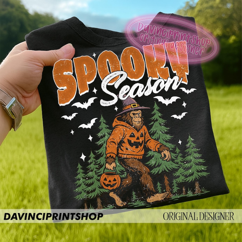 May include: Black t-shirt with the words "SPOOKY Season" in orange and white. The shirt features a Bigfoot character wearing a pumpkin-themed hoodie and holding a jack-o'-lantern. Bats and pine trees complete the Halloween design.