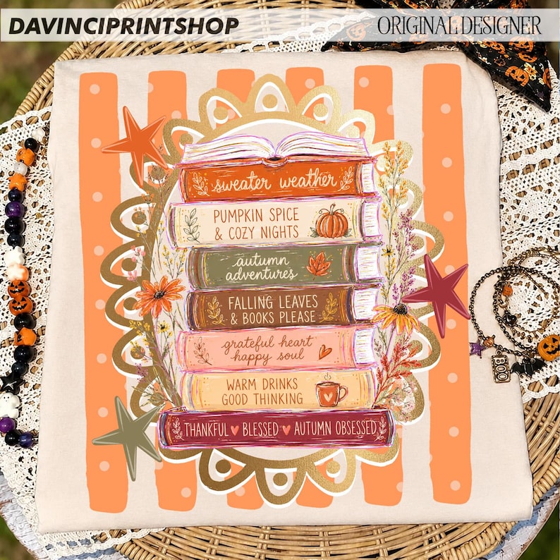 K&ouml;nnte beinhalten: Ein beiges T-Shirt mit einem Stapel bunter B&uuml;cher und herbstlichen Texten wie sweater weather, pumpkin spice und autumn adventures. Das Design ist von einem dekorativen goldenen Rand, orangefarbenen Streifen und Sternen umgeben.